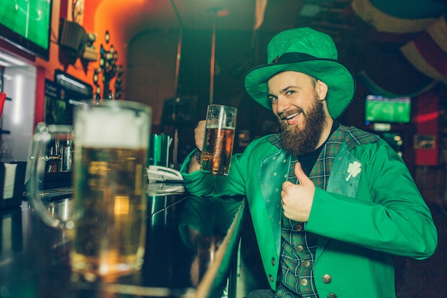 Jovem barbudo feliz e animado em traje de São Patrício sentar no balcão de bar no pub e segurar a caneca de cerveja. Ele levantou o polegar grande. Outra caneca de cerveja fica perto da câmera.