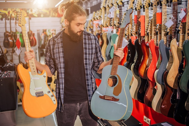 Jovem barbudo ficar e segurar guitarras elétricas e acústicas. ele olha para o azul. hipster jovem sozinho na sala cheia de guitarras e equipamentos diferentes.