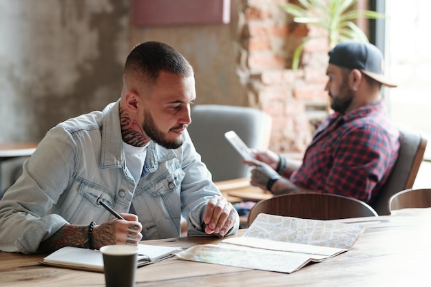 Jovem barbudo focado sentado à mesa em um café e escolhendo lugares no mapa de papel enquanto planejava as férias