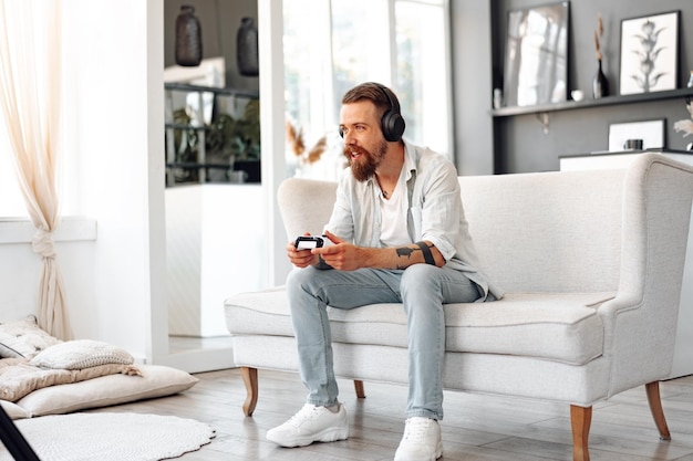 Foto jovem barbudo jogando videogame enquanto está sentado no sofá em casa