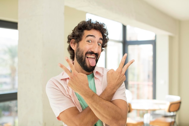 jovem barbudo louco sorrindo e parecendo feliz, amigável e satisfeito, gesticulando vitória ou paz