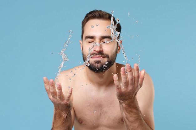 Jovem barbudo nu de 20 anos cercado por gotas de água lavando o rosto isolado no retrato de estúdio de fundo pastel azul. Conceito de procedimentos cosméticos de saúde para cuidados com a pele Mock up espaço de cópia