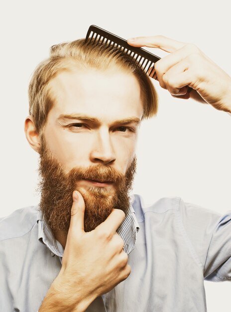 Jovem barbudo penteando o cabelo e olhando para a câmera em pé contra um fundo cinza claro