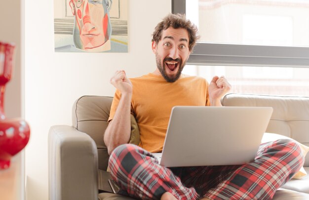 Jovem barbudo se sentindo chocado, animado e feliz, rindo e comemorando o sucesso, dizendo uau e sentado com um laptop