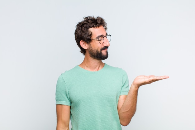 Jovem barbudo se sentindo feliz e sorrindo casualmente, olhando para um objeto ou conceito seguro na mão ao lado