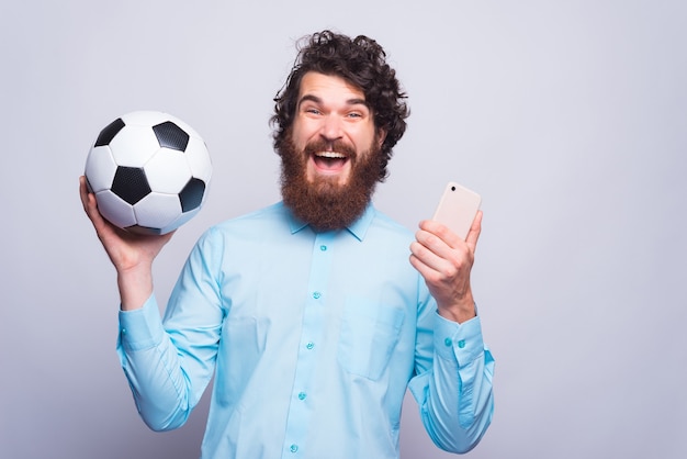 Jovem barbudo segurando um celular e uma bola de futebol