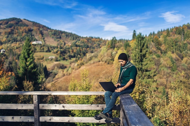 Jovem barbudo senta-se no corrimão e trabalha no laptop. hipster freelancer de férias nas montanhas.