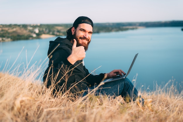 Jovem barbudo sentado em um campo com seu computador sorrindo e segurando um polegar perto de um rio