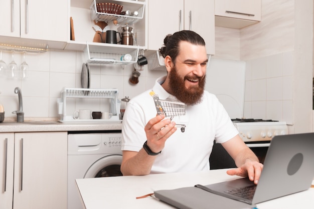 Jovem barbudo sentado na cozinha em frente ao laptop e comprando algo online