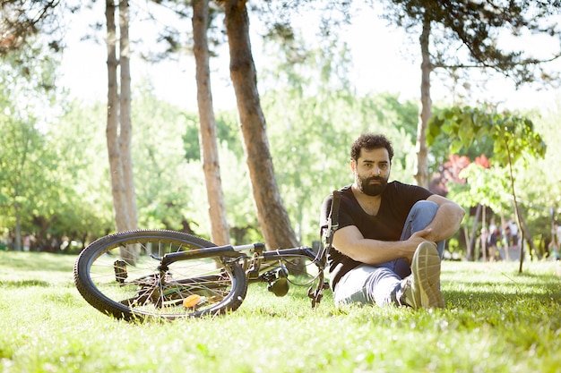 Jovem barbudo sentado na grama do parque ao lado de sua bicicleta