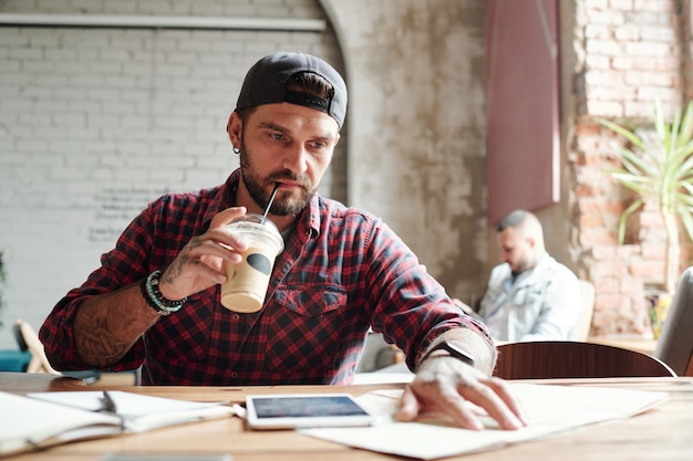 Jovem barbudo sério com boné bebendo café frio e verificando lugares no mapa de papel em um café