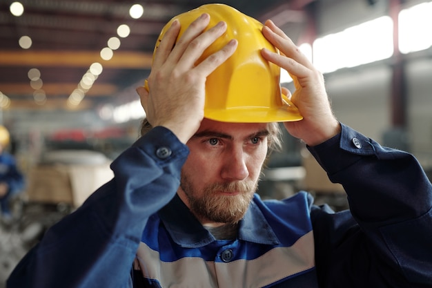 Foto jovem barbudo sério em pé na fábrica e colocando capacete de segurança na cabeça enquanto se prepara para o trabalho