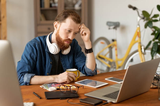 Jovem barbudo sério focado no conceito de design, sentado à mesa e olhando para o desenho na tela do laptop