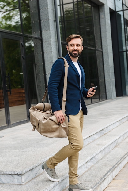 Jovem barbudo sorridente atraente vestindo jaqueta, caminhando ao ar livre na rua, carregando uma bolsa, segurando um telefone celular
