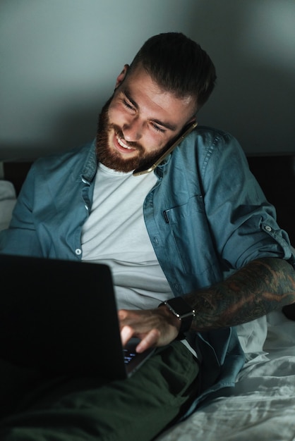 Jovem barbudo sorridente, deitado na cama em casa à noite, falando no celular enquanto trabalha no laptop