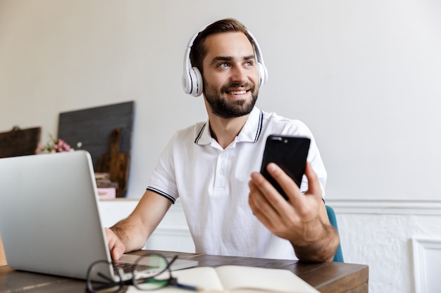 Jovem barbudo sorridente e bonito sentado à mesa em casa, usando um laptop, um telefone celular