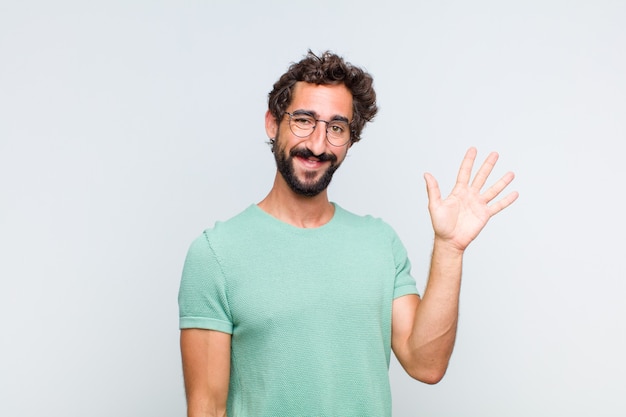 Jovem barbudo sorrindo feliz e alegre, acenando com a mão, dando as boas-vindas e cumprimentando você ou dizendo adeus