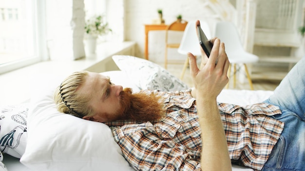 Jovem barbudo usando computador tablet deitado na cama no quarto em casa
