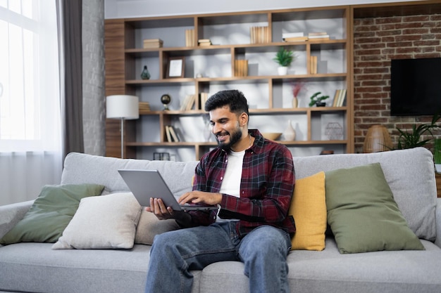 Jovem barbudo usando laptop sentado no sofá em casa bonitão fazendo videochamada