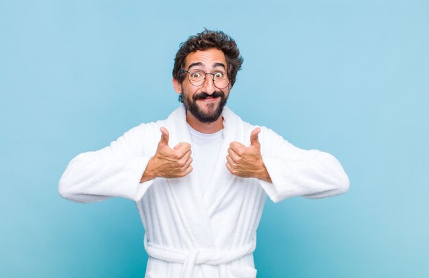 Jovem barbudo vestindo um roupão de banho sorrindo e parecendo feliz, positivo, confiante e bem-sucedido, com os dois polegares para cima