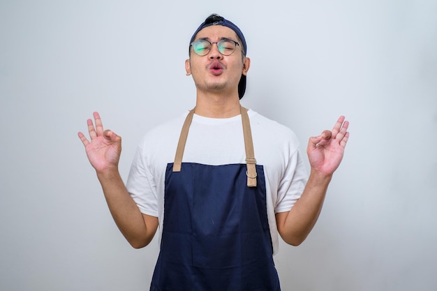 Jovem barista asiático bonito vestindo avental em pé relaxe e sorrindo com os olhos fechados fazendo gesto de meditação com os dedos Conceito de ioga