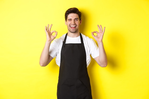 Jovem barista de avental preto mostrando sinais de ok e sorrindo, garantia de qualidade na cafeteria, em pé sobre fundo amarelo.