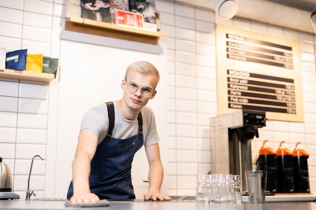Jovem barista ou trabalhador de um café aconchegante contemporâneo preparando seu local de trabalho ou mesas para clientes pela manhã