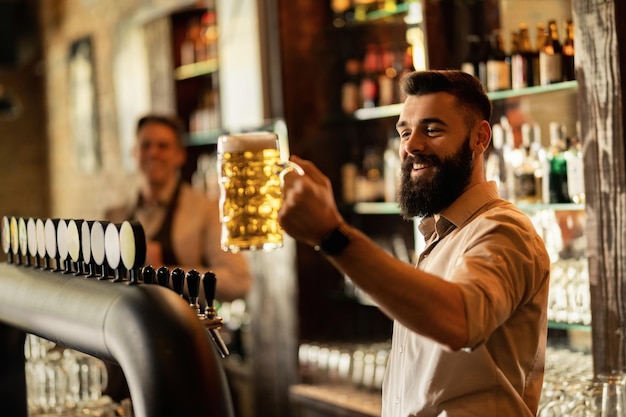 Jovem barman feliz segurando um copo de cerveja enquanto trabalhava em um pub