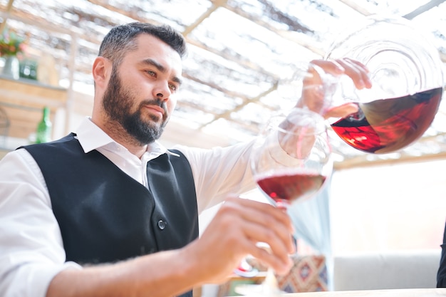 Jovem barman ou sommelier barbudo servindo vinho tinto em uma taça de vinho enquanto trabalhava na adega ou no bar