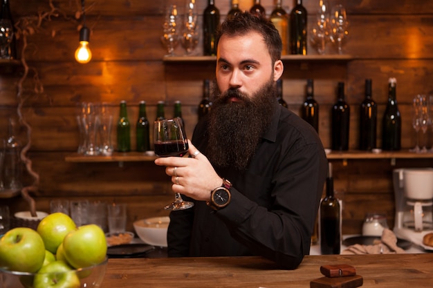 Jovem barman tatuado com grande barba, degustando uma taça de vinho. pub moderno.