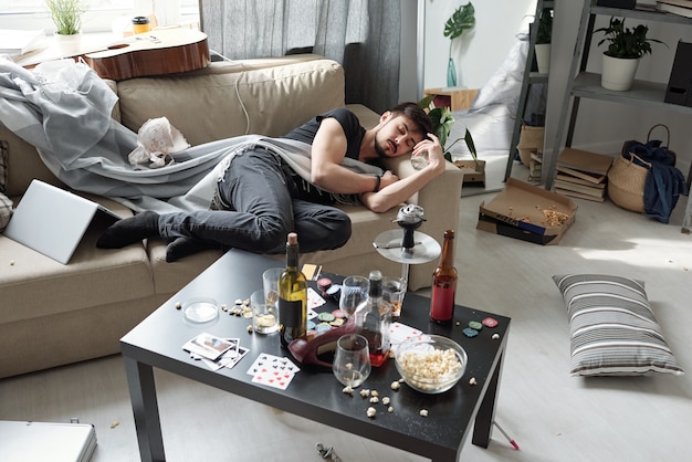 Jovem bêbado dormindo no sofá entre coisas bagunçadas em uma sala suja com uma garrafa de álcool na mesa