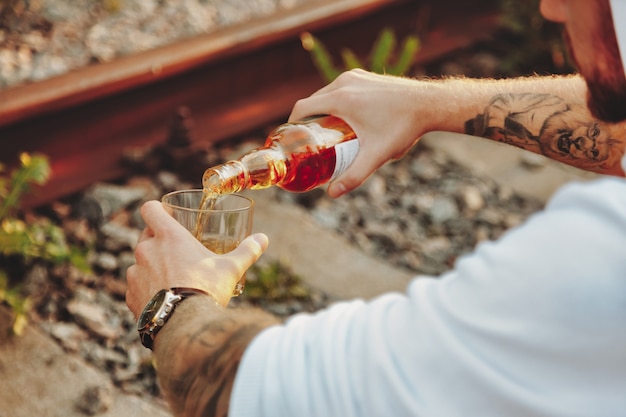 Jovem bebe álcool em uma ferrovia abandonada na zona rural ao pôr do sol. Conceito de tristeza, apatia, depressão ou estilo de vida incorreto. Copie o espaço