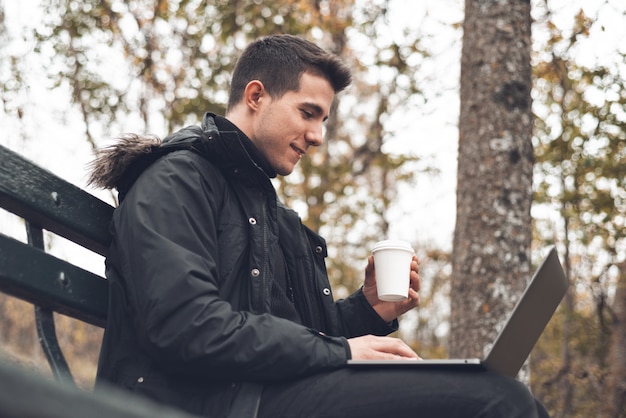 Jovem bebendo café com laptop no outono park ao ar livre. Homem com laptop e café no parque outono sob folhagem de outono.