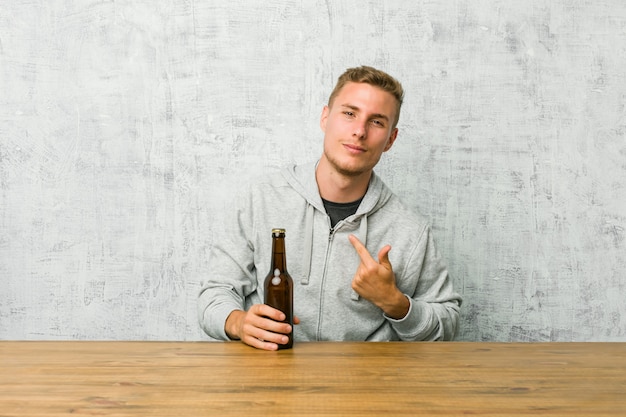 Jovem bebendo uma cerveja, uma mesa apontando com o dedo como se estivesse convidando para se aproximar.