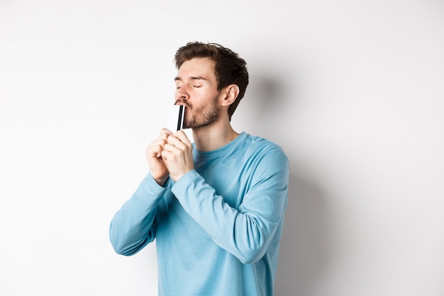 Foto jovem beijando o cartão de crédito de plástico com o rosto satisfeito, em pé sobre um fundo branco em uma camisa azul casual.