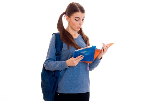 Jovem bela estudante com mochila lendo um livro e posando isolada na parede branca em estúdio