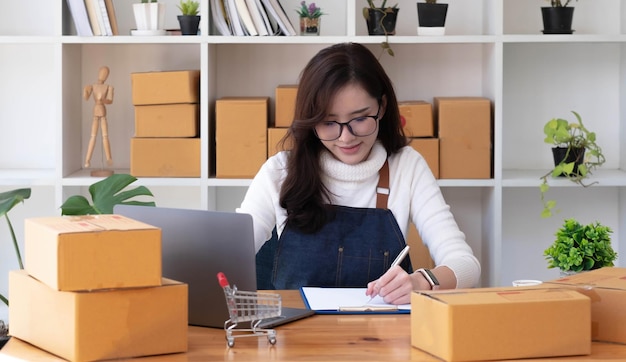 Jovem bela feliz mulher de negócios asiática proprietária de SME on-line usando laptop receber encomenda de cliente com embalagem de caixa de encomenda em seu escritório em casa inicial vendedor de negócios on-line e entrega