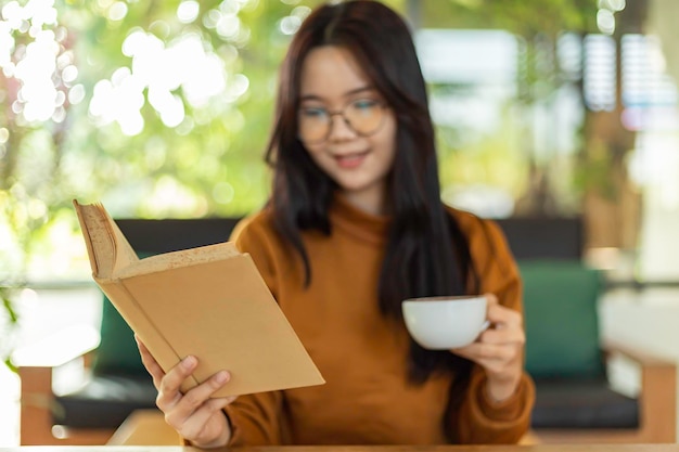 Jovem bela mulher asiática segurando caneca e lendo um livro enquanto relaxa durante o trabalho em casa