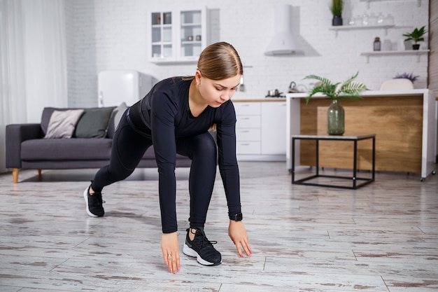 Jovem bela mulher atlética em legging preta e top fazendo exercícios de aquecimento em casa. Estilo de vida saudável. Garota com roupas de ginástica. A mulher pratica esportes em casa.