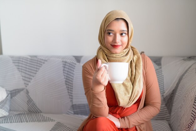 Jovem bela mulher muçulmana com café pensando na sala de estar em casa