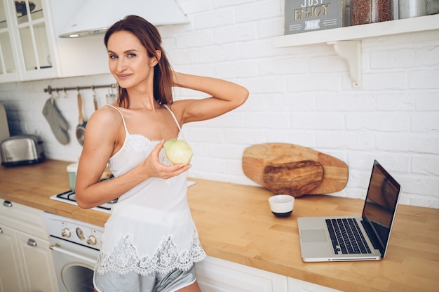 Jovem bela mulher sorridente, comendo uma maçã na cozinha e usando o computador portátil