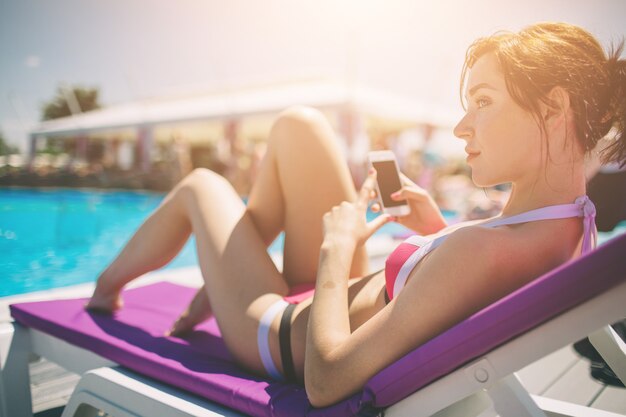 Jovem bela mulher sorridente em biquíni na piscina quente no resort e falar no celular.