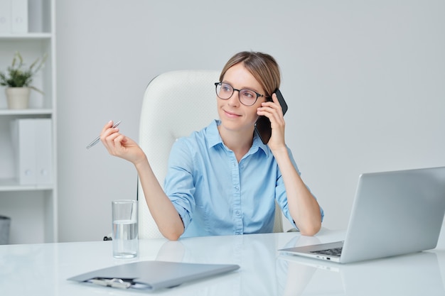 Jovem bem-sucedida e confiante chefe de empresa com o telefone perto da orelha, sentada perto da mesa no escritório e ouvindo um dos clientes