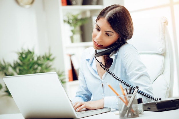 Jovem bem sucedida trabalhando em sua mesa no escritório. Ela está usando o telefone e trabalhando no laptop.