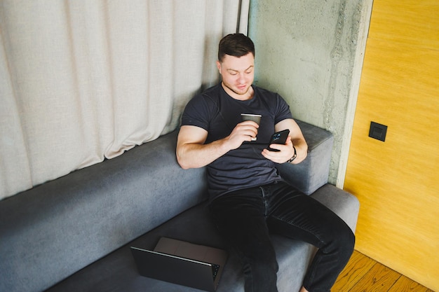 Jovem bem-sucedido em camiseta sentado no sofá com telefone e tomando café trabalhando remotamente durante a inicialização como freelancer olhando para laptop e sorrindo