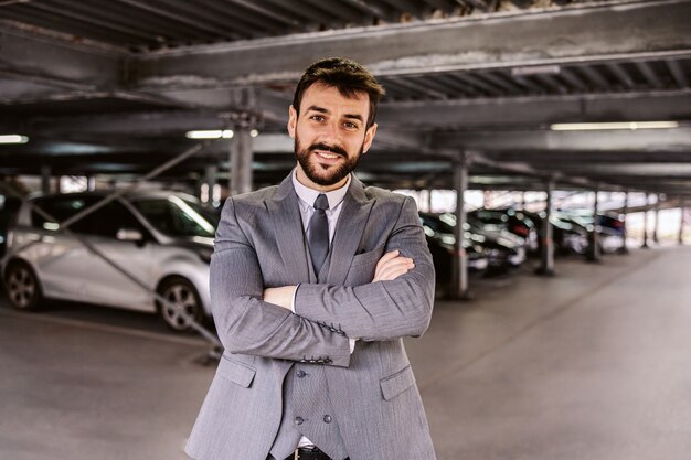 Jovem bem sucedido negociante de automóveis barbudo de terno em pé no estacionamento com os braços cruzados