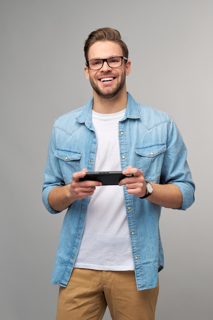 Jovem, bem-vindo, jogando videogame portátil em pé sobre uma parede cinza