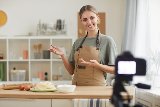 Jovem blogueira de avental ensinando seus seguidores a cozinhar online na cozinha doméstica