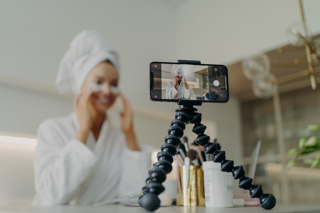 Jovem blogueira de beleza feliz vestindo roupão de banho, gravando um vídeo tutorial para cuidados com a pele. blog sobre o uso de adesivos cosméticos sob os olhos para tratar rugas e olheiras, foco na tela do smartphone