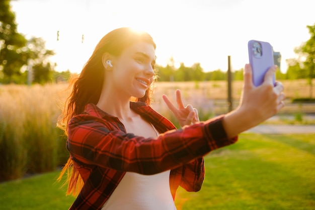 Foto jovem blogueira ruiva tira uma selfie e mostra paz ao pôr do sol usando um smartphone